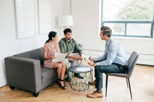 A real estate agent sitting across from a couple on a sofa, explaining real estate commissions and holding documents in a bright, modern living room setting.