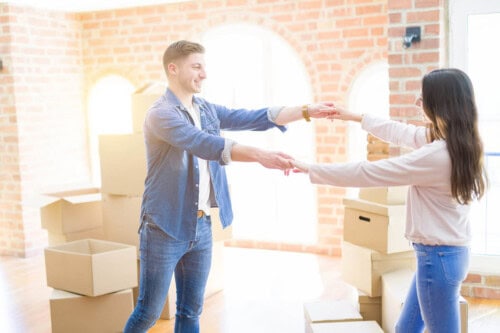 A happy couple dances in a bright, brick-walled room surrounded by moving boxes. They are likely celebrating after purchasing a new home, possibly after navigating multiple offers on the house.