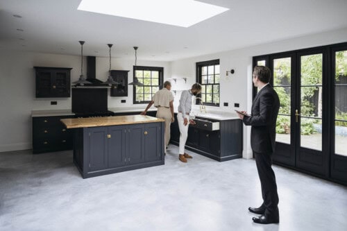 A real estate agent stands in a modern kitchen observing two potential homebuyers inspecting the cabinetry and sink, possibly negotiating repairs after a home inspection.