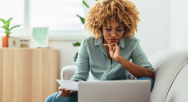 A Black woman wearing glasses, focused on a laptop screen while holding a sheet of paper in her hand. 